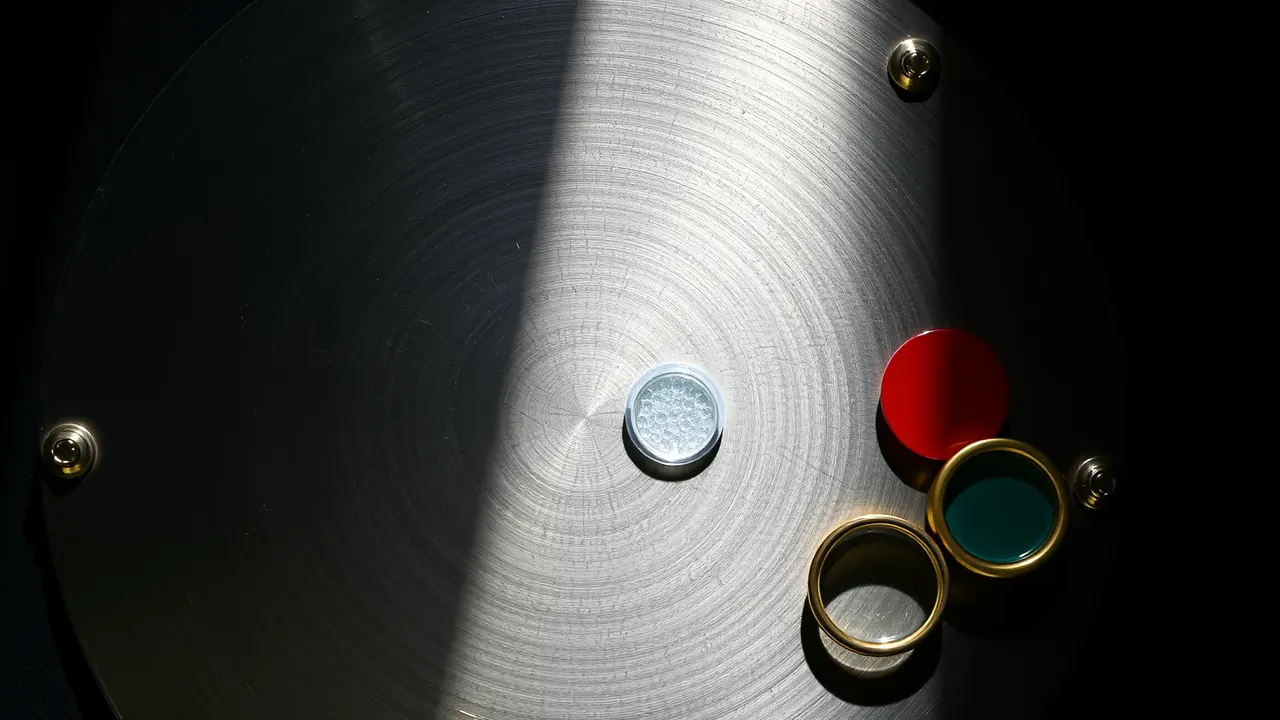 A large brushed steel plate with a perfectly smooth unmarked face and four small round rivets at its corners, seen close from slightly above, a brass magnifying loupe resting on its lower edge, and three flat glossy enamel discs in a short row beside it with a clear gap between each, emerald green, deep violet and scarlet red.